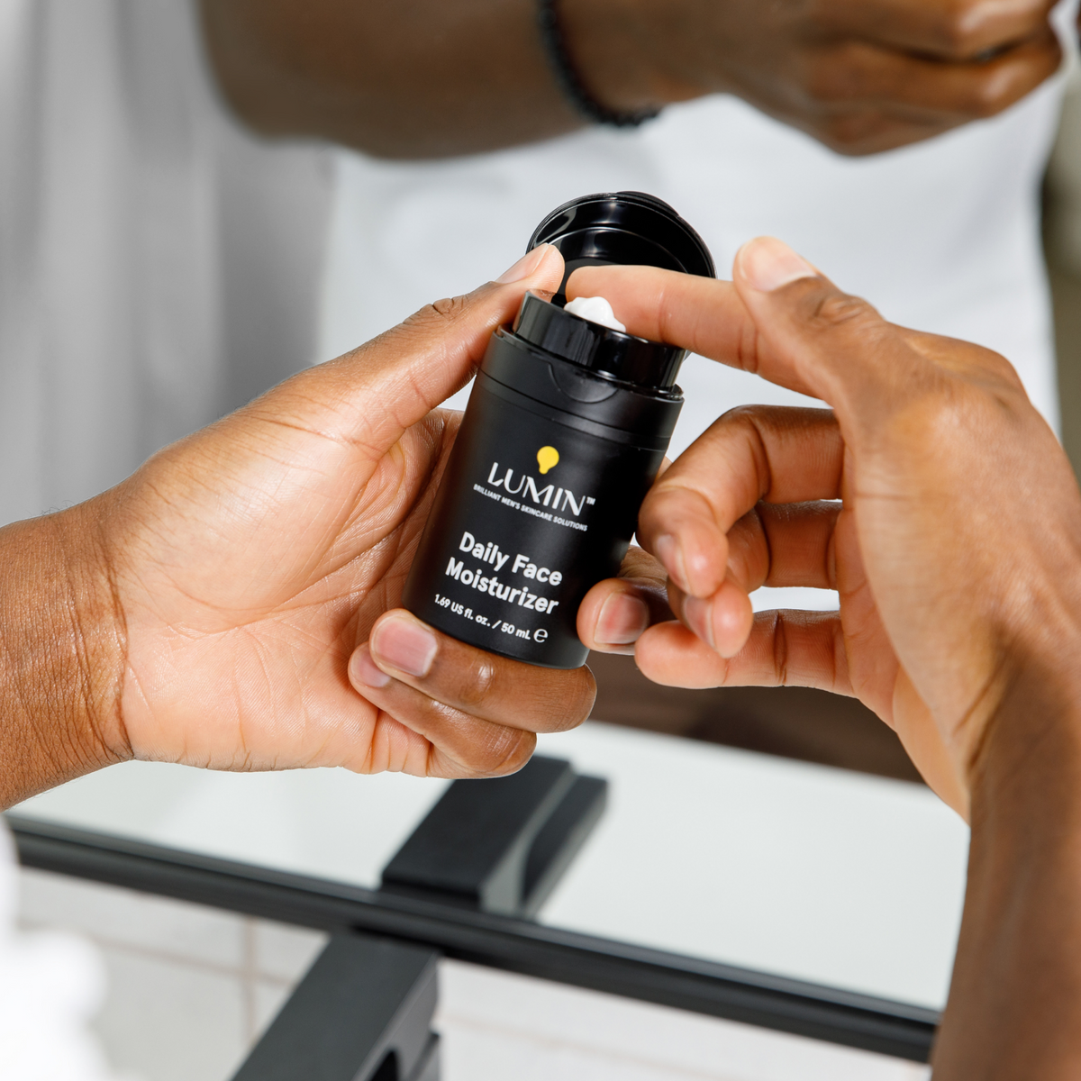 Black luxury skincare jar labeled "LILAVIN Daily Face Renewing Cream" held by two dark-skinned hands near a bathroom mirror, skincare routine.