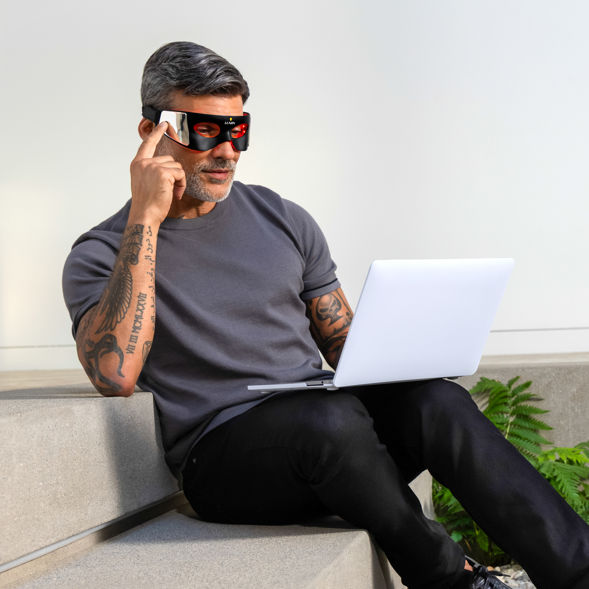 Man with tattoos wearing black sunglasses using laptop while sitting on concrete bench in modern indoor space