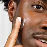 Close-up of dark-skinned man applying white moisturizing cream to smooth facial skin for skincare routine.