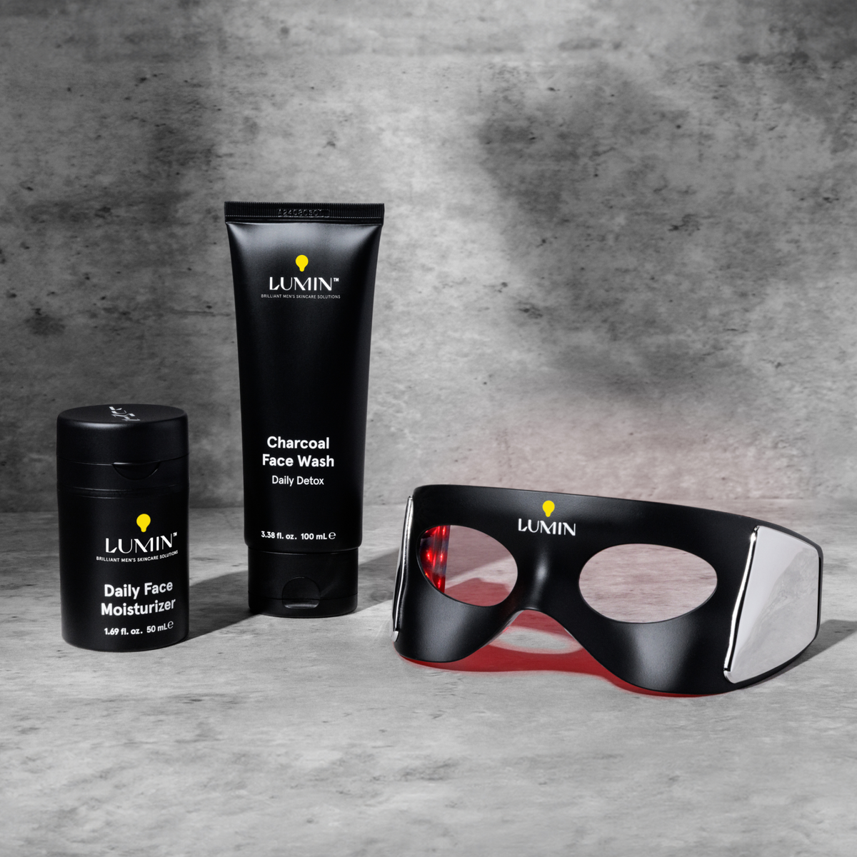 Lumin men's skincare set with Daily Face Moisturizer, Charcoal Face Wash, and LED eye massage mask on gray background.
