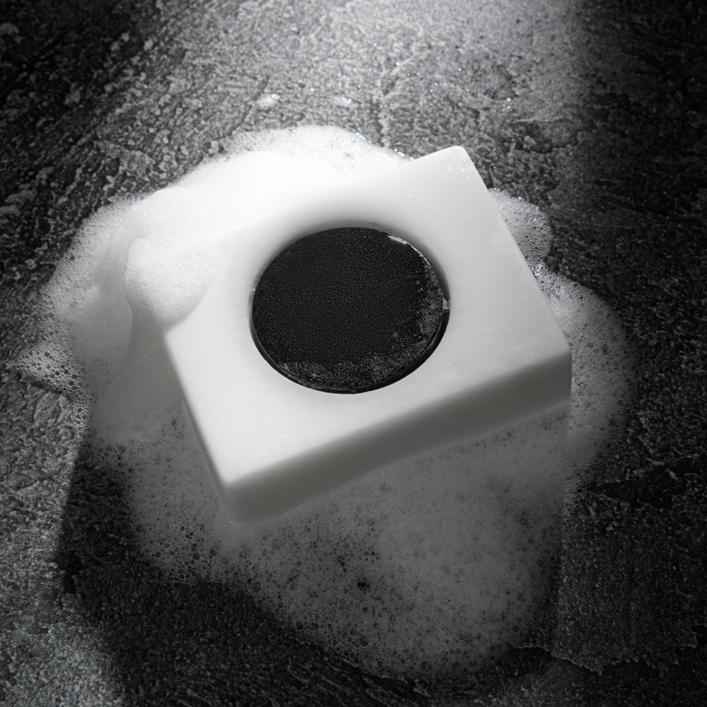 White soap bar with black circular center on wet dark surface surrounded by soap bubbles, handwashing and hygiene concept.