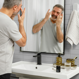Middle-aged man applying anti-aging cream in bathroom mirror beside skincare products and modern faucet on white sink.