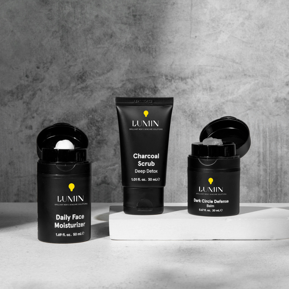 Lumin men's black skincare set with Daily Face Moisturizer, Charcoal Scrub, and Dark Circle Defense Balm on modern gray background