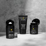 Lumin men's black skincare set with Daily Face Moisturizer, Charcoal Scrub, and Dark Circle Defense Balm on modern gray background