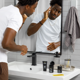 Man in white t-shirt grooming skin in bathroom mirror with skincare products, towel, and comb on sink counter