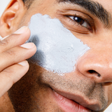 Man applying gray clay facial mask on cheek for skincare, grooming, and men's beauty routine close-up