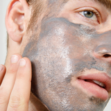Man applying gray clay face mask on cheek with fingers, skincare routine, facial treatment for men, close-up.