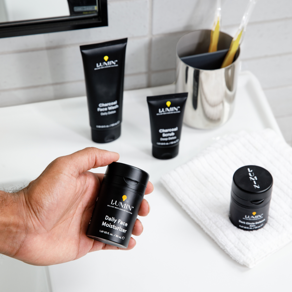 Hand holding Lumin Daily Face Moisturizer with Charcoal Face Wash, Scrub, and dark circle defense on white bathroom sink.