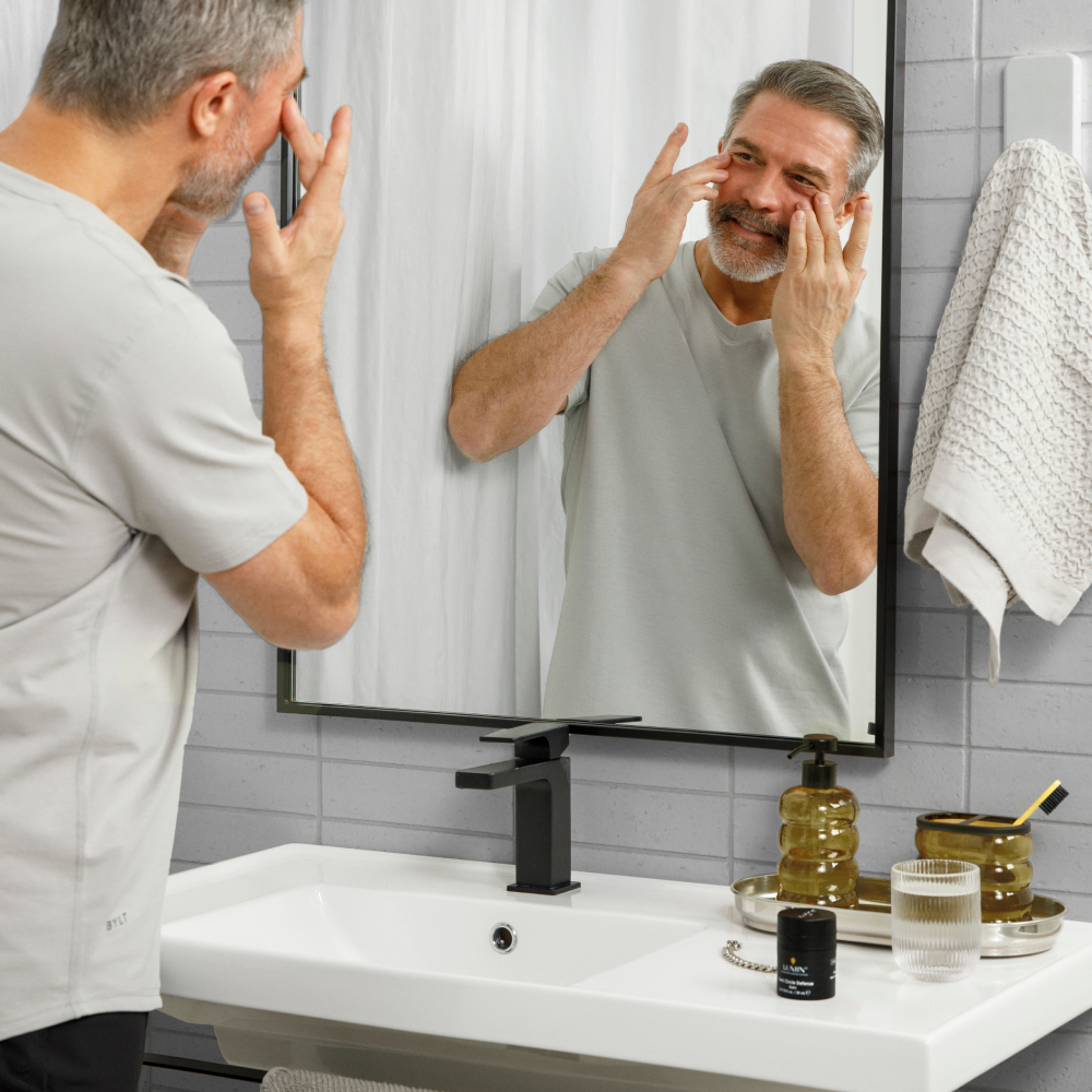 Middle-aged man applying anti-aging cream to face in modern bathroom with skincare products on sink.