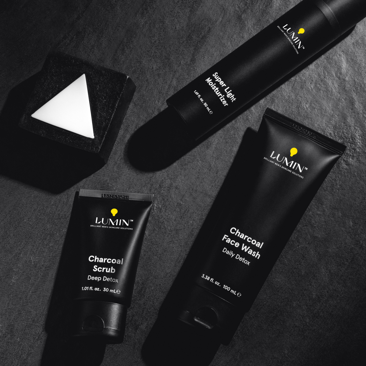 Lumin men's skincare set with charcoal scrub, face wash, super light moisturizer, and a geometric black soap bar on dark surface.