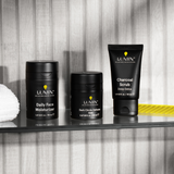 Men's skincare set with Lumin Daily Face Moisturizer, Dark Circle Defense Balm, and Charcoal Scrub on glass shelf.