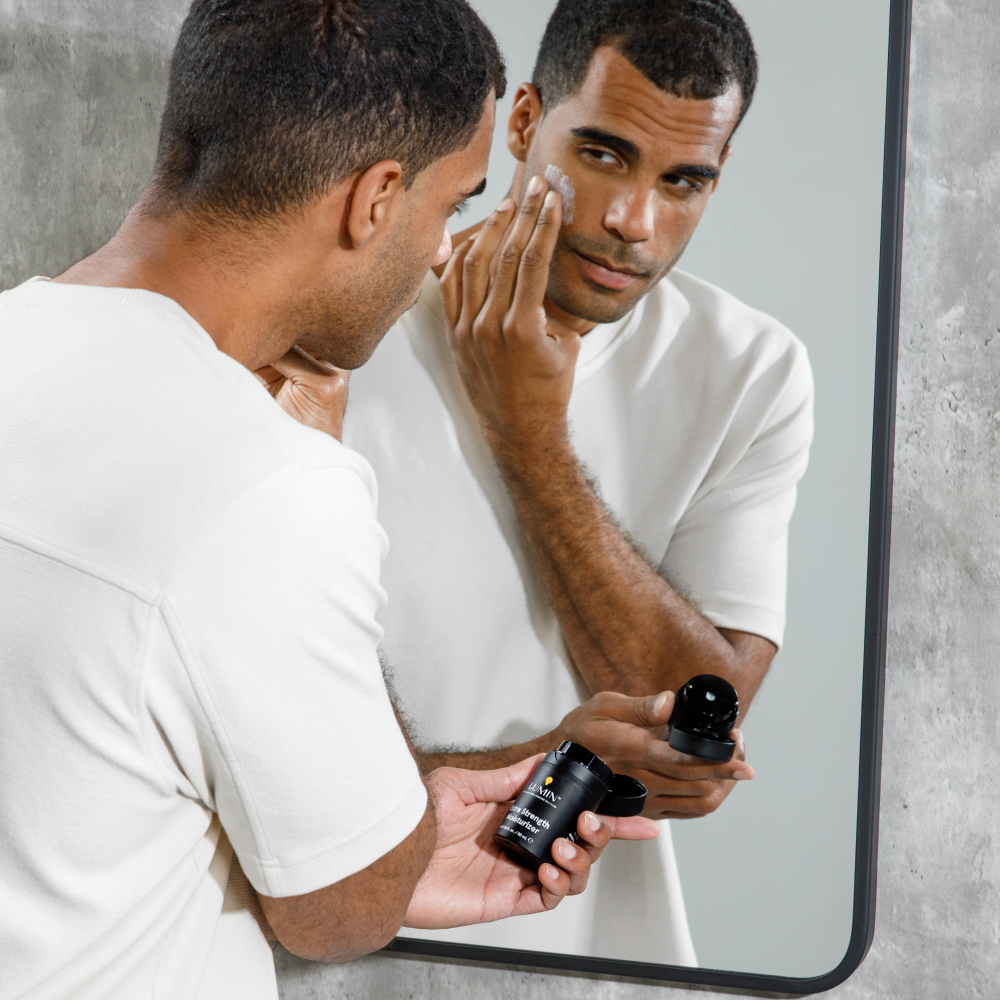 Young man applying moisturizer cream from black jar while looking in bathroom mirror, skincare routine for men