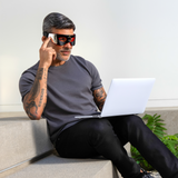 Man with tattoos wearing smart glasses and casual outfit, using laptop while sitting on modern concrete bench with plant nearby