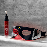 Red light therapy eye mask with adjustable strap and companion eye serum bottle on gray textured background, wellness skincare device.