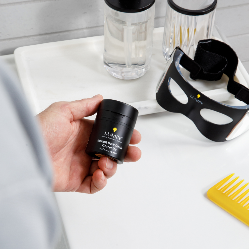 Hand holding Lumin Instant Dark Circle Corrector next to black light therapy eye mask, clear skincare bottles, and yellow comb on white surface