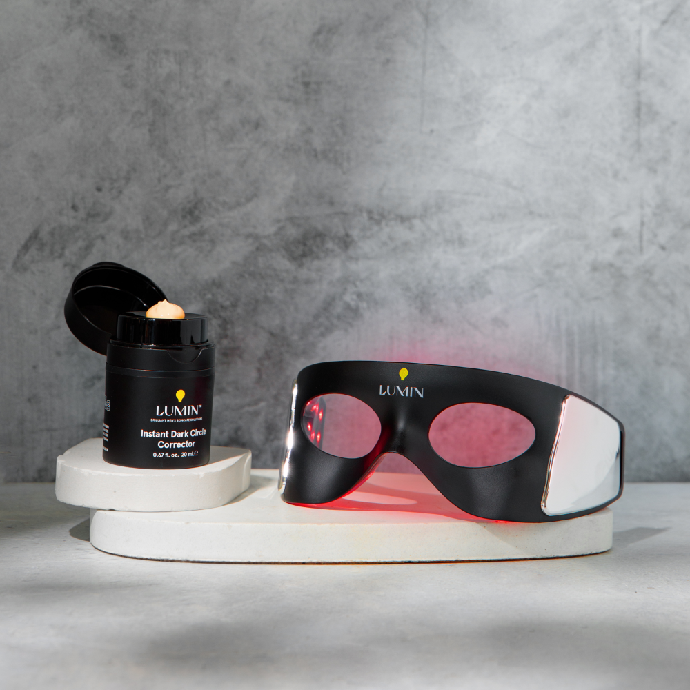 Lumin Instant Dark Circle Corrector cream and black LED light therapy eye mask on white display stand with gray background.