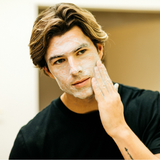 Young man with styled hair applying white facial cleanser cream in bathroom mirror, skincare routine for men.