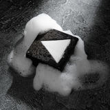 Black triangular bar soap with white triangle detail surrounded by white foam bubbles on textured dark surface