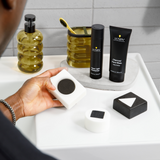 Hand holding white soap bar with black circle, near charcoal face wash, moisturizer, yellow toothbrush, and soap dispenser on bathroom sink.
