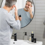 Middle-aged man applying face serum in bathroom with skincare products, including charcoal face wash and moisturizer, on the sink.