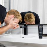 Man washing face with Lumin Charcoal Face Wash near modern black faucet and white sink in bathroom mirror reflection