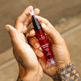 Hydrating red lip oil with applicator held by tattooed hands, natural background, skincare and beauty product.