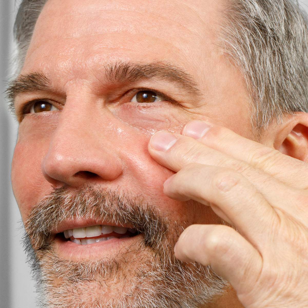 Middle-aged man with gray beard applying skincare cream to face, demonstrating facial skincare routine for men.