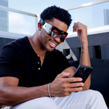 Man wearing futuristic Lumin eye mask, smiling and using smartphone, casual black shirt and white pants, outdoor seating.