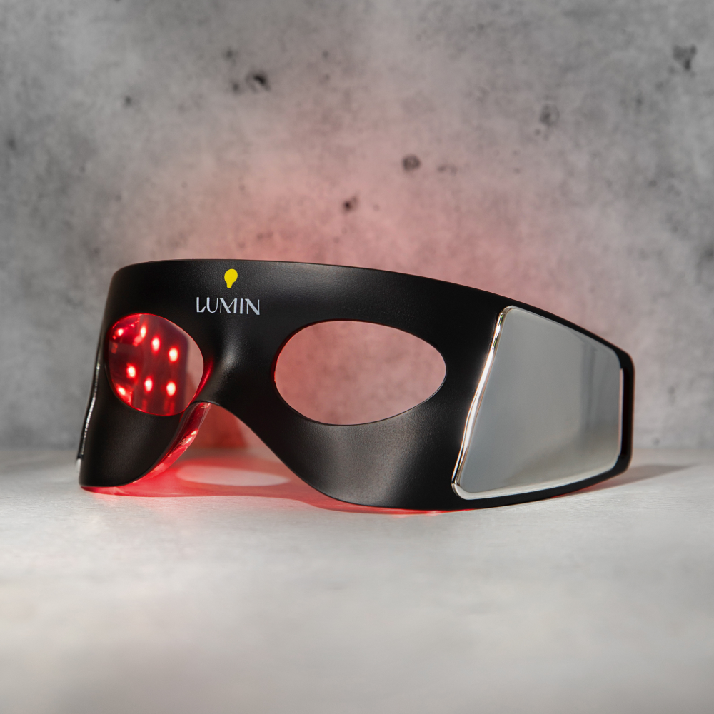 Black Lumin LED light therapy mask with red LED lights glowing inside and sleek silver side panels on gray surface.