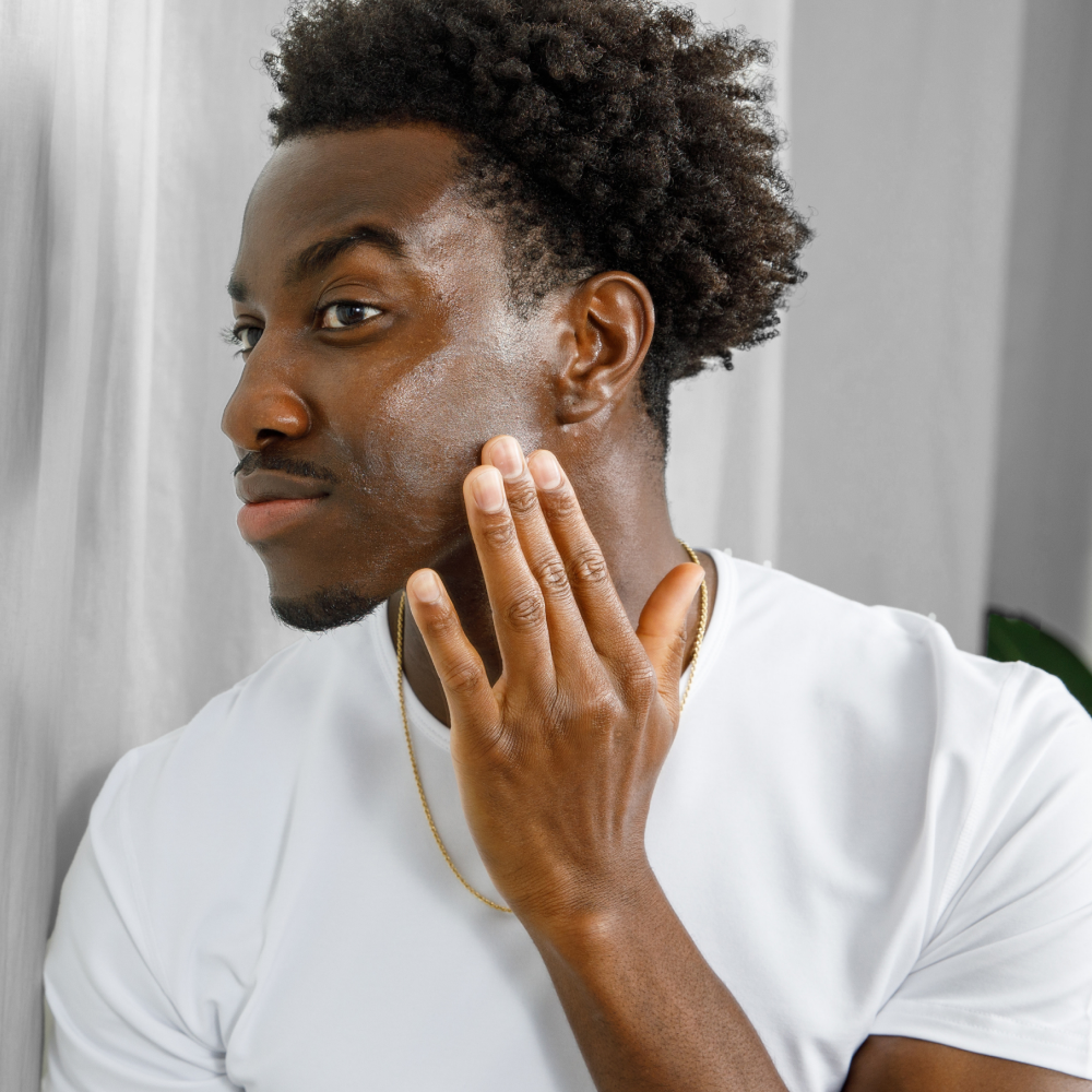 Young Black man applying facial moisturizer for skincare routine, wearing white shirt and gold chain necklace indoors.