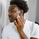 Young Black man applying facial moisturizer for skincare routine, wearing white shirt and gold chain necklace indoors.