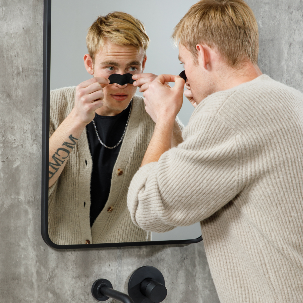 Young man applying black nose pore strip in bathroom mirror, skincare routine, men's grooming, clear skin treatment.