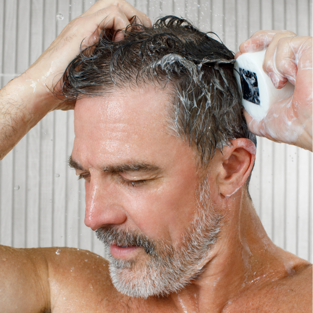Middle-aged man washing gray-streaked hair with shampoo bar under running water in tiled shower