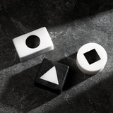 Black and white geometric blocks with cutout shapes including circle, triangle, and square on textured dark surface.