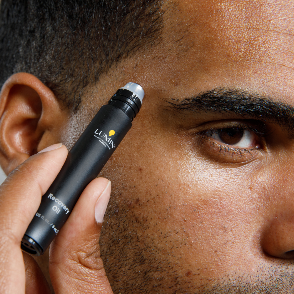Lumin Recovery Oil roller applied near eye on man's textured skin, skincare product for dark circles and puffiness.