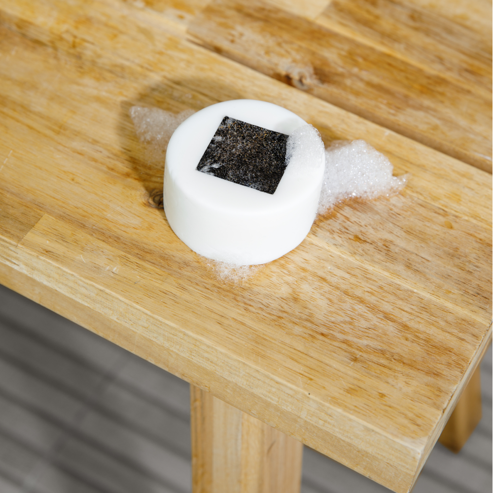 Round white soap bar with a black square center and foam bubbles on a wooden table surface.