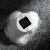Black charcoal soap bar surrounded by white creamy foam on textured dark gray surface, skincare cleansing product.