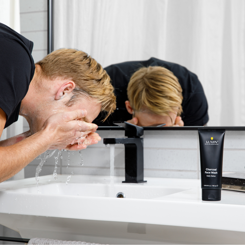 Man washing face with water in modern bathroom sink, Lumin charcoal face wash tube visible for daily detox skincare.