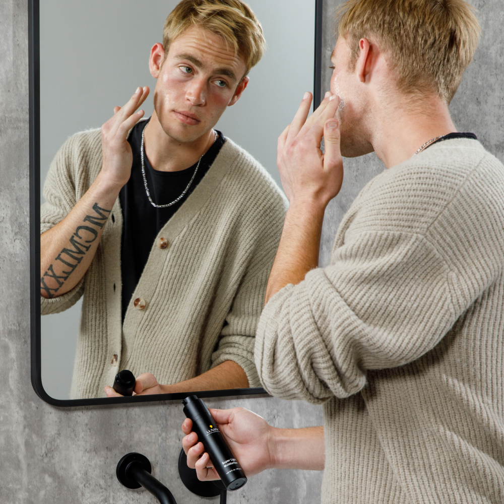 Young man in beige cardigan applying moisturizing face serum in front of bathroom mirror, skincare routine for men.