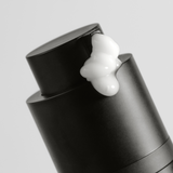 Black pump bottle dispensing thick white moisturizer cream against a plain gray background.